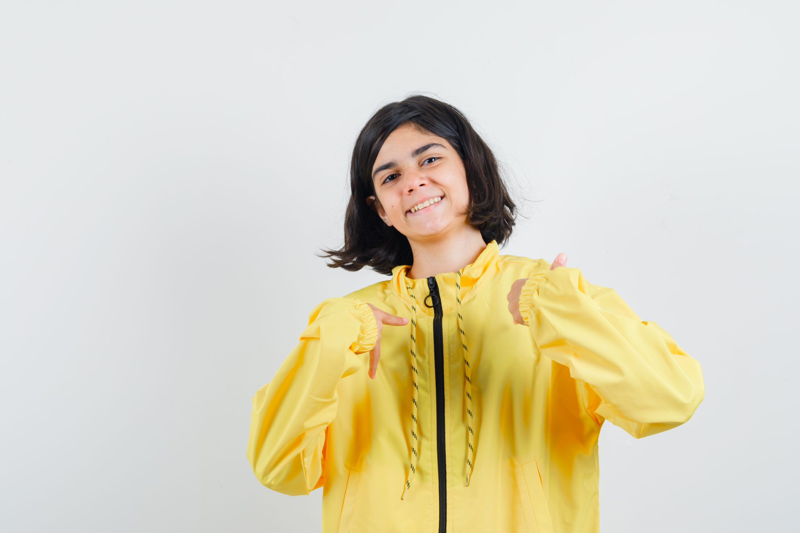 young girl pointing herself with index fingers in yellow bomber jacket and looking happy , front view.